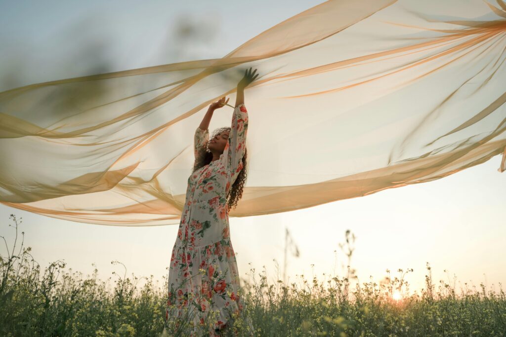 A woman stands in a field of flowers, arms raised, against a sunset backdrop.