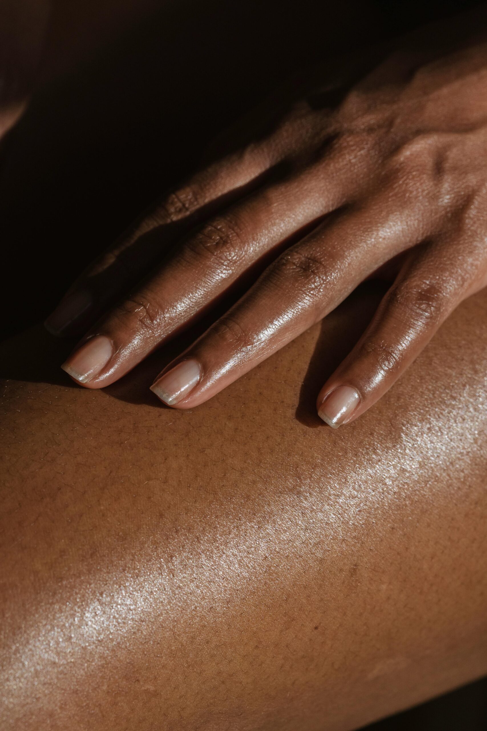 Crop anonymous African American female touching bare leg with hand while sitting on black background in room with bright sunlight