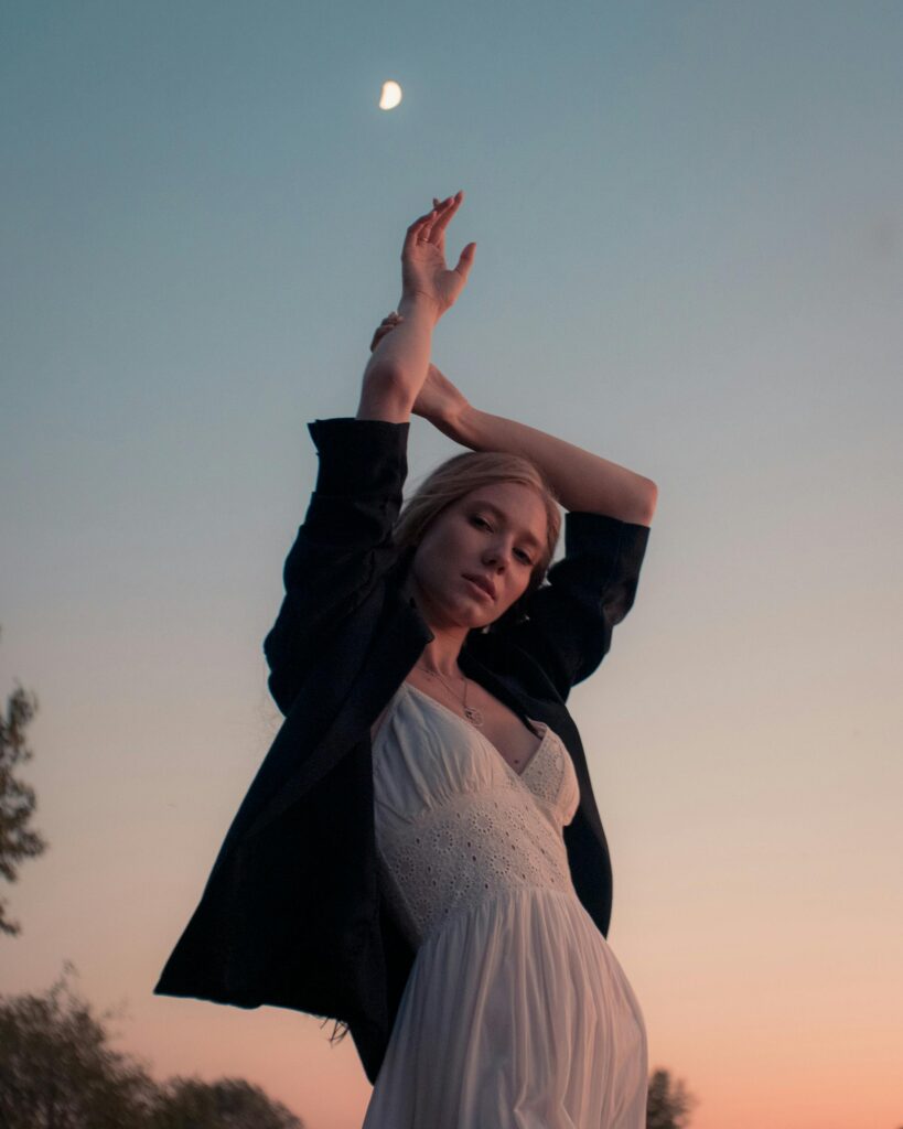 A peaceful young woman in a white dress poses gracefully at dusk with a crescent moon in the sky above.